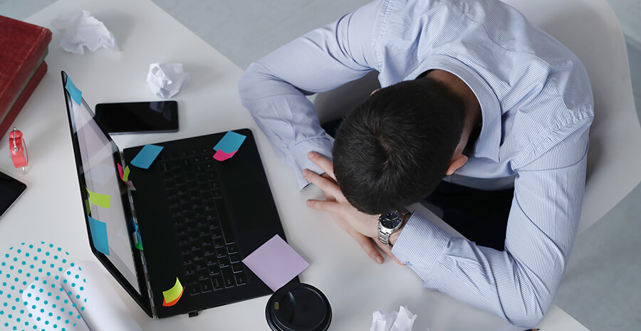 imagem de um homem cansado em frente ao notebook com vários papéis amassados na mesa para representar a Síndrome de Burnout